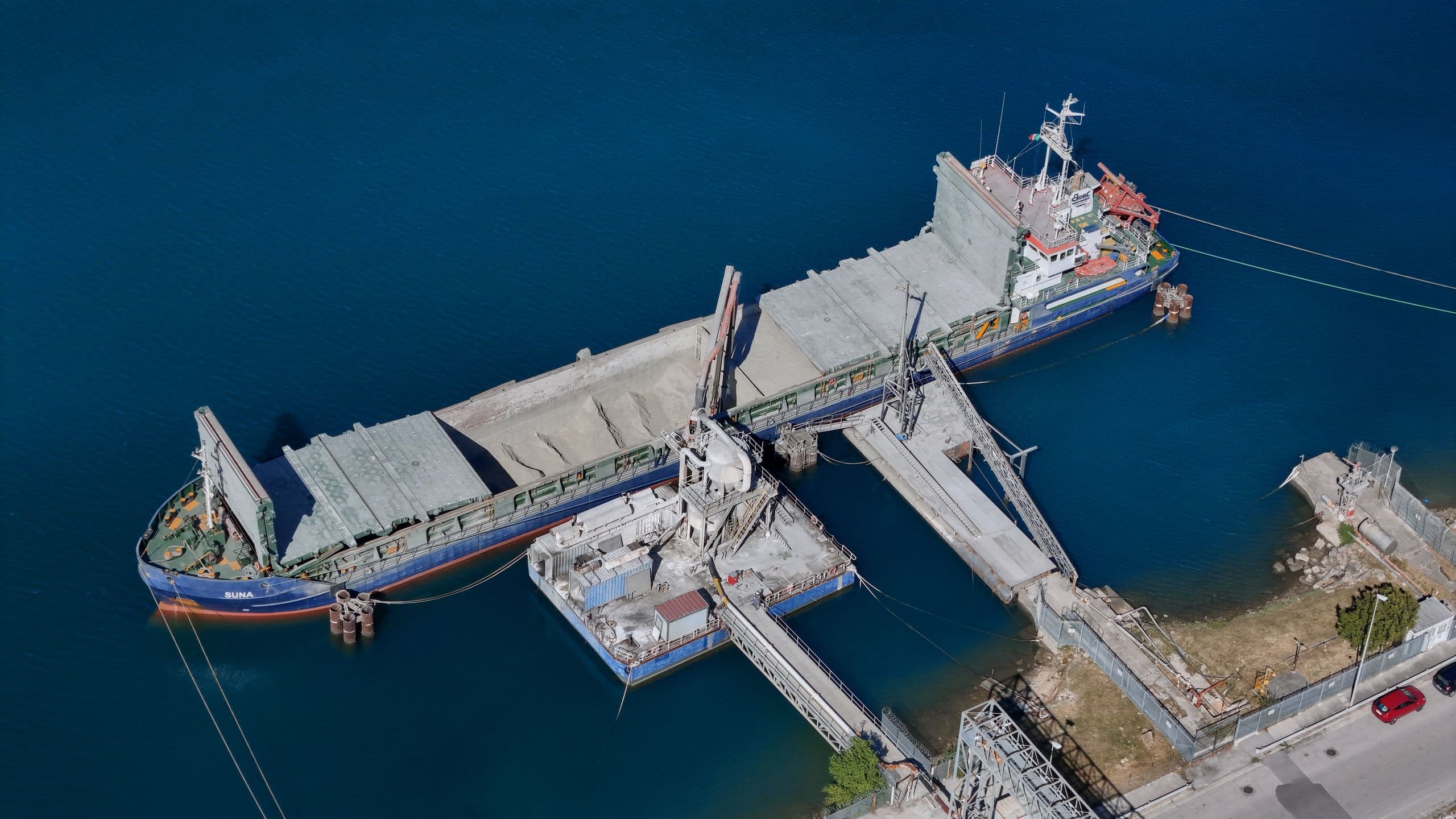 A bulk carrier moored alongside a jetty, loading dry cargo into its open holds through a shore conveyor.